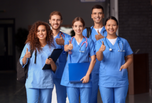 A group of medical students in blue scrubs smiling and giving thumbs up, showing enthusiasm and readiness for their profession.